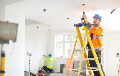 contractor on a latter wearing construction gear installing a light fixture on the ceiling and another in the background doing an installation