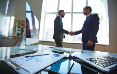 two men shaking hands in an open room with stats on a clipboard along with a laptop