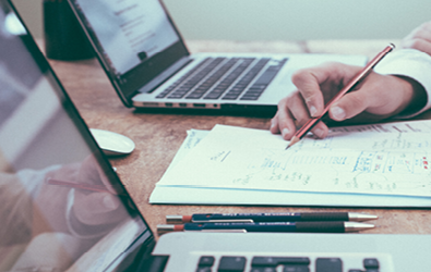 a hand holding a pencil to paper in between two laptops